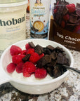 A white bowl filled with Greek yogurt, fresh raspberries and pieces of dark chocolate cereal, with Chobani Greek Yogurt, Miss Mary's Sinless Syrup bottle, and Cereal visible in the background on a granite countertop.