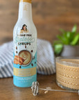 Miss Mary's Almond Biscotti coffee syrup on wood table with glass mug of coffee and frother against blurred background.