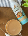 Almond Biscotti Sinless Syrup on a striped towel with a latte, frother, and plant on wood table.
