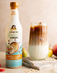 Apple crisp syrup bottle with an iced apple crisp oatmilk macchiato and apples on a beige background