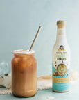 A bottle of Cinnamon Dolce coffee syrup stands next to a mason jar filled with iced coffee and a metal straw, on a light blue surface with a soft blue background.