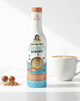 Bottle of sugar-free hazelnut Sinless Syrup next to a cup of coffee on a white background