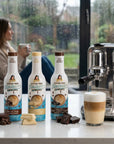 Chocolate Lovers trio of sugar-free Sinless coffee syrups on a counter with a coffee machine and a cup of coffee.