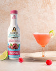 Bottle of Sugar Free Sinless Syrups next to a cocktail with lime and raspberry garnishes on a neutral background