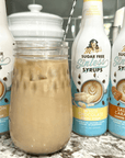 An iced coffee drink sits on a countertop, surrounded by Miss Mary's Sugar Free Sinless Syrups in Cinnamon Dolce, White Chocolate, and Salted Caramel flavors.