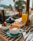 A styled breakfast table scene with coffee, orange juice, a bottle of sugar-free Salted Caramel syrup, fruit, toast, and utensils on a woven placemat, with an outdoor balcony and blurred background.