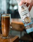 Hand holding a bottle of sugar-free salted dark chocolate syrup next to a tall espresso drink.