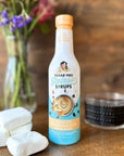Toasted Marshmallow Sinless Syrup bottle with large marshmallows on a wood surface with coffee and flowers blurred in the background