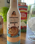 Bottle of Mrs. Mary's Sugar Free Sinless Syrups Toasted Marshmallow flavor on a table with a can, carton, and glass.