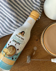 White Chocolate Sinless Syrup on wood table with frother and latte, striped towel.