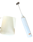 A stainless steel frothing pitcher next to an electric frother wand with a white handle and a USB charger.