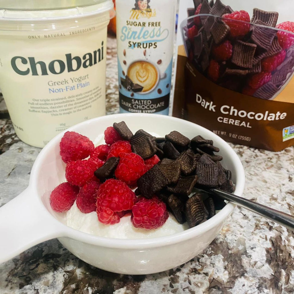 A white bowl filled with Greek yogurt, fresh raspberries and pieces of dark chocolate cereal, with Chobani Greek Yogurt, Miss Mary's Sinless Syrup bottle, and Cereal visible in the background on a granite countertop.