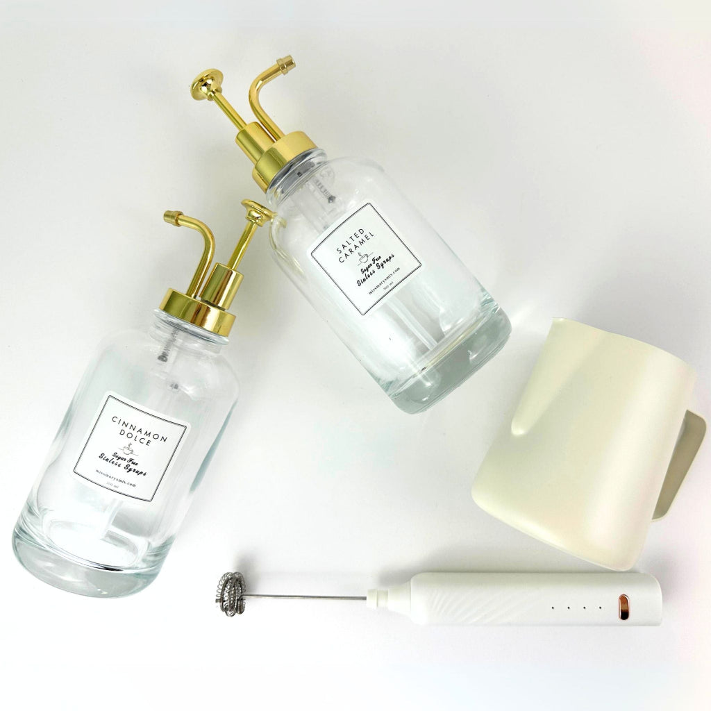 Two glass bottles with gold dispensers, a white electric frother, and a white mug on a light gray background.