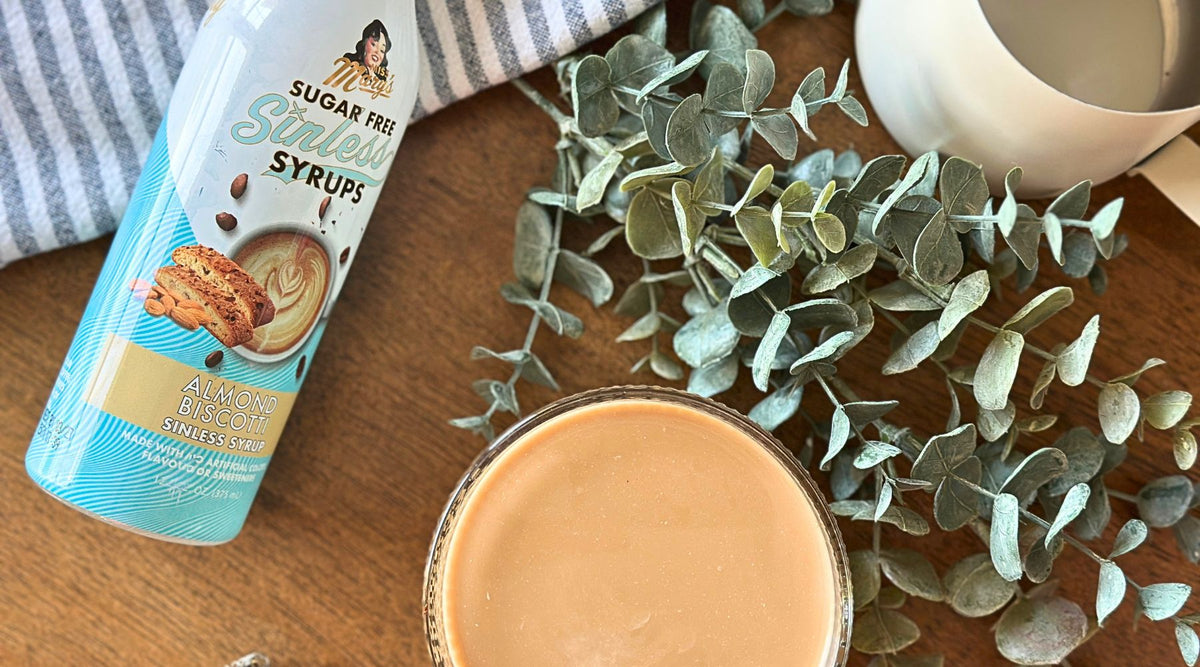 A bottle of Almond Biscotti sugar-free Sinless syrup on a wooden table beside a glass of coffee with milk, a white pitcher, striped towel, and green eucalyptus leaves.