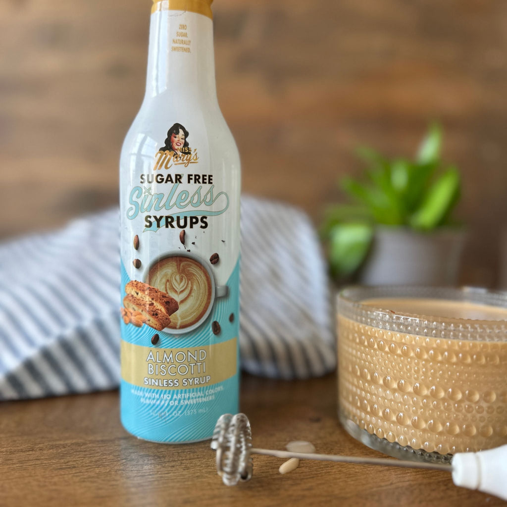 Miss Mary's Almond Biscotti coffee syrup on wood table with glass mug of coffee and frother against blurred background.