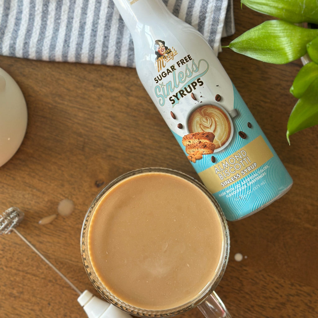 Almond Biscotti Sinless Syrup on a striped towel with a latte, frother, and plant on wood table.