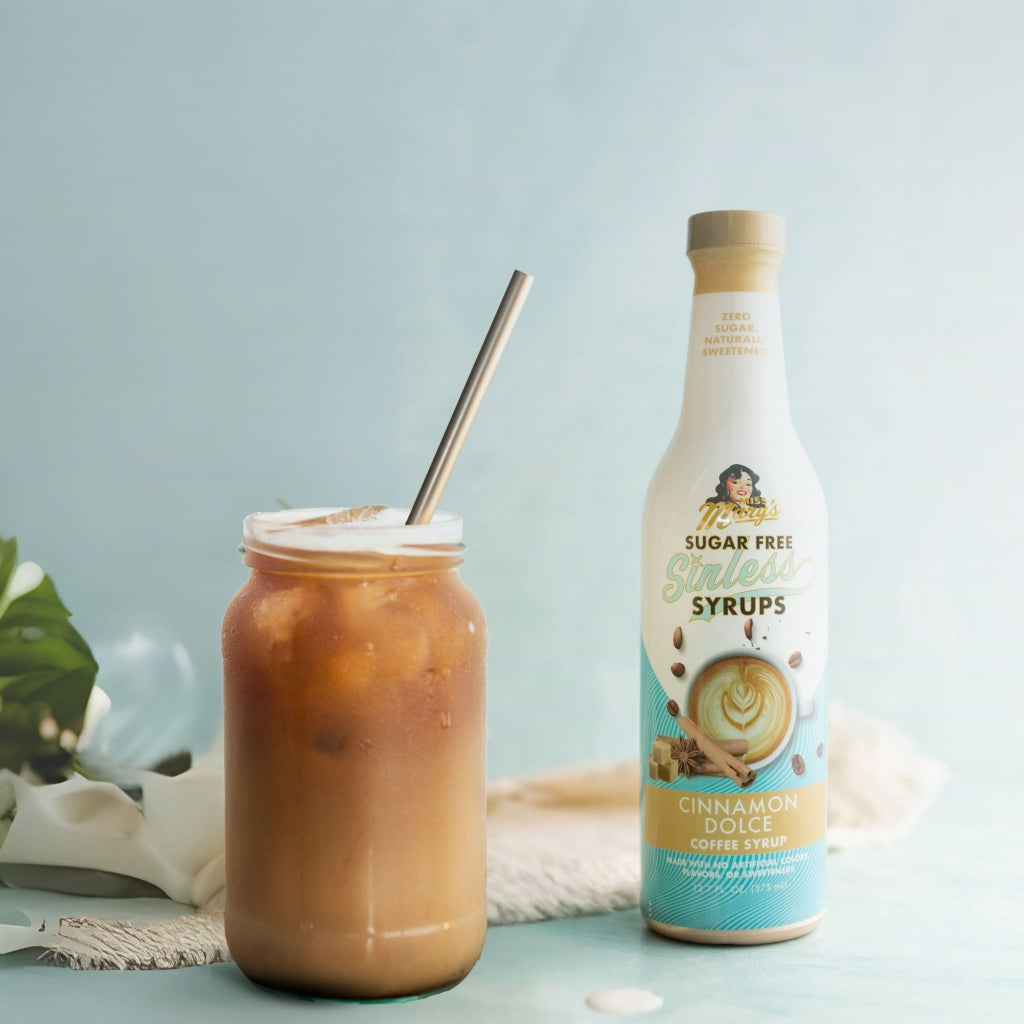 A bottle of Cinnamon Dolce coffee syrup stands next to a mason jar filled with iced coffee and a metal straw, on a light blue surface with a soft blue background.