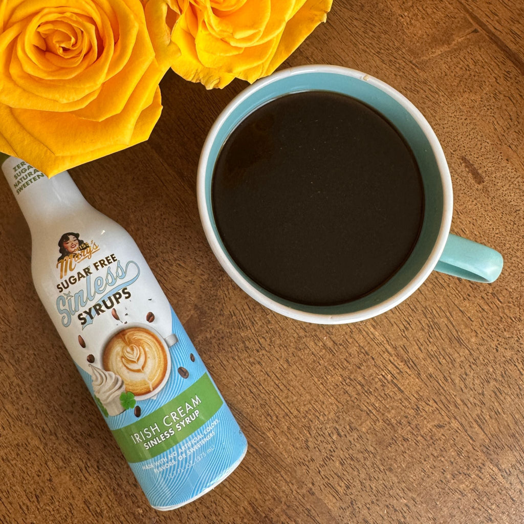 Miss Mary's Irish Cream sugar-free coffee syrup against a wood table with a blue coffee mug and yellow roses
