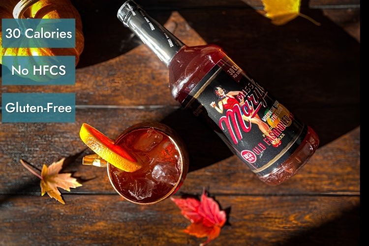A bottle of Miss Mary's Old Fashioned Mix on wood table with cocktail glass and orange slice garnish. Three blue text boxes on the left read '30 Calories', 'No HFCS', and 'Gluten-Free'.