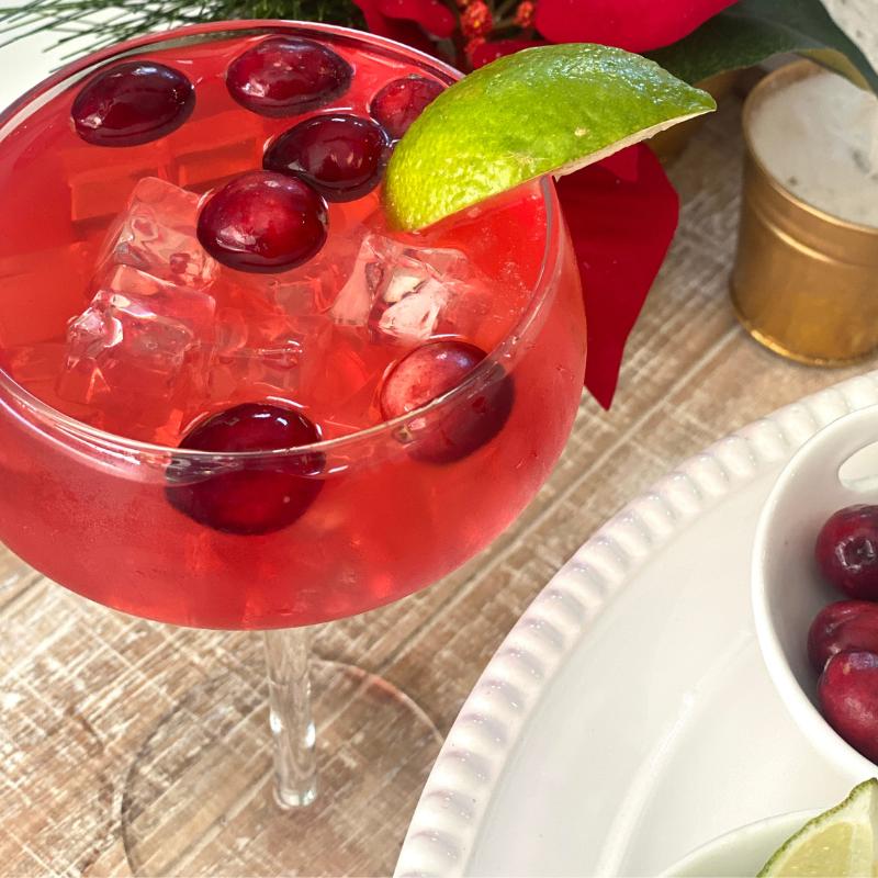 Red cocktail with cranberries and a lime wedge in a glass, placed on a textured surface.