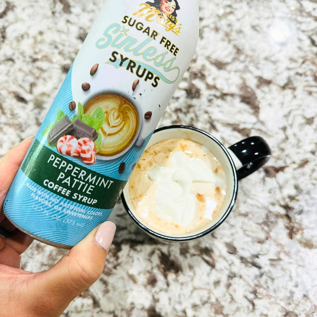 A hand holds a bottle of Miss Mary's Sugar Free Sinless Peppermint Pattie coffee syrup next to a black mug filled with a hot chocolate drink topped with whipped cream, set on a speckled granite countertop.