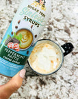 A hand holds a bottle of Miss Mary's Sugar Free Sinless Peppermint Pattie coffee syrup next to a black mug filled with a hot chocolate drink topped with whipped cream, set on a speckled granite countertop.