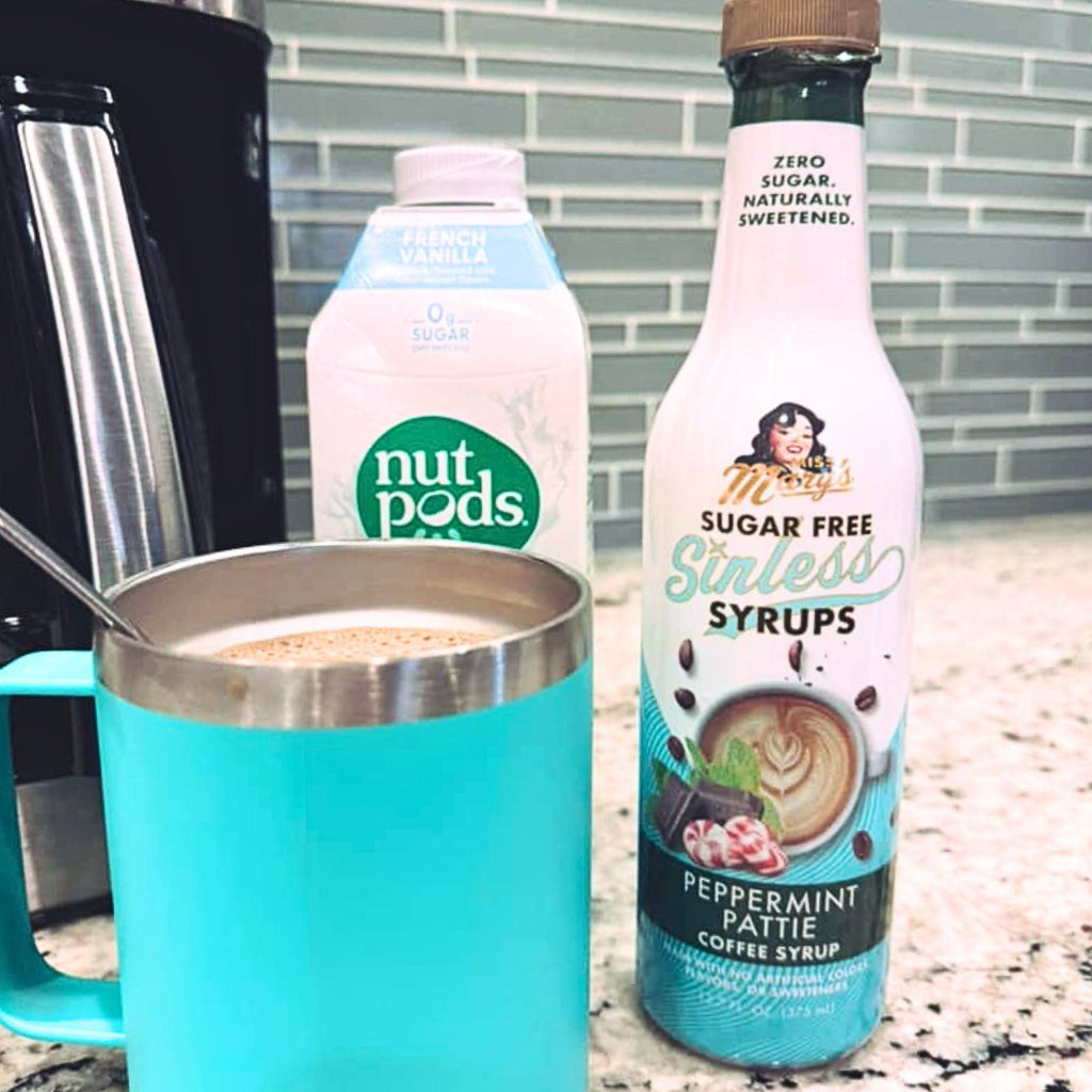 A turquoise insulated coffee mug filled with coffee sits on a granite countertop next to a bottle of Mary's Sugar Free Sinless Syrups Peppermint Pattie Coffee Syrup and a carton of Nut Pods Vanilla.