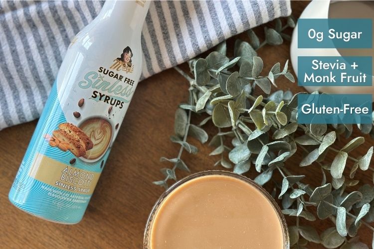 Almond Biscotti Sinless Syrup bottle with latte, striped towel, and greenery with text "0g Sugar, Stevia + Monk Fruit, Gluten-Free"