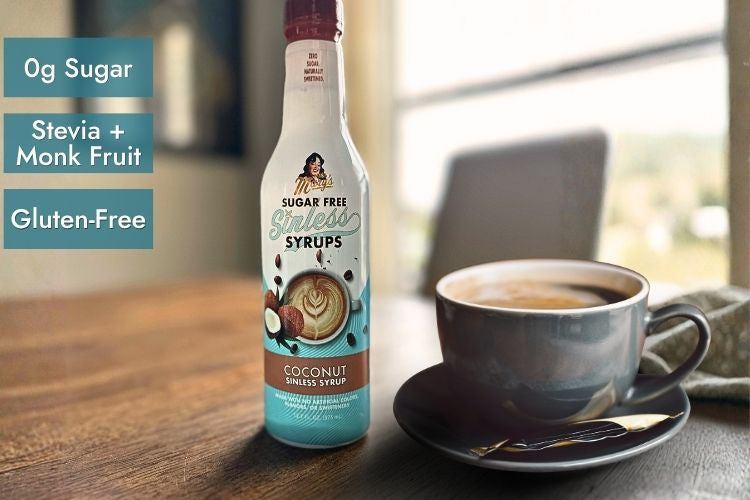 A bottle of sugar-free coconut syrup stands on a wooden table beside a cup of coffee on a saucer. Text on the left reads '0g Sugar', 'Stevia + Monk Fruit', and 'Gluten-Free'.