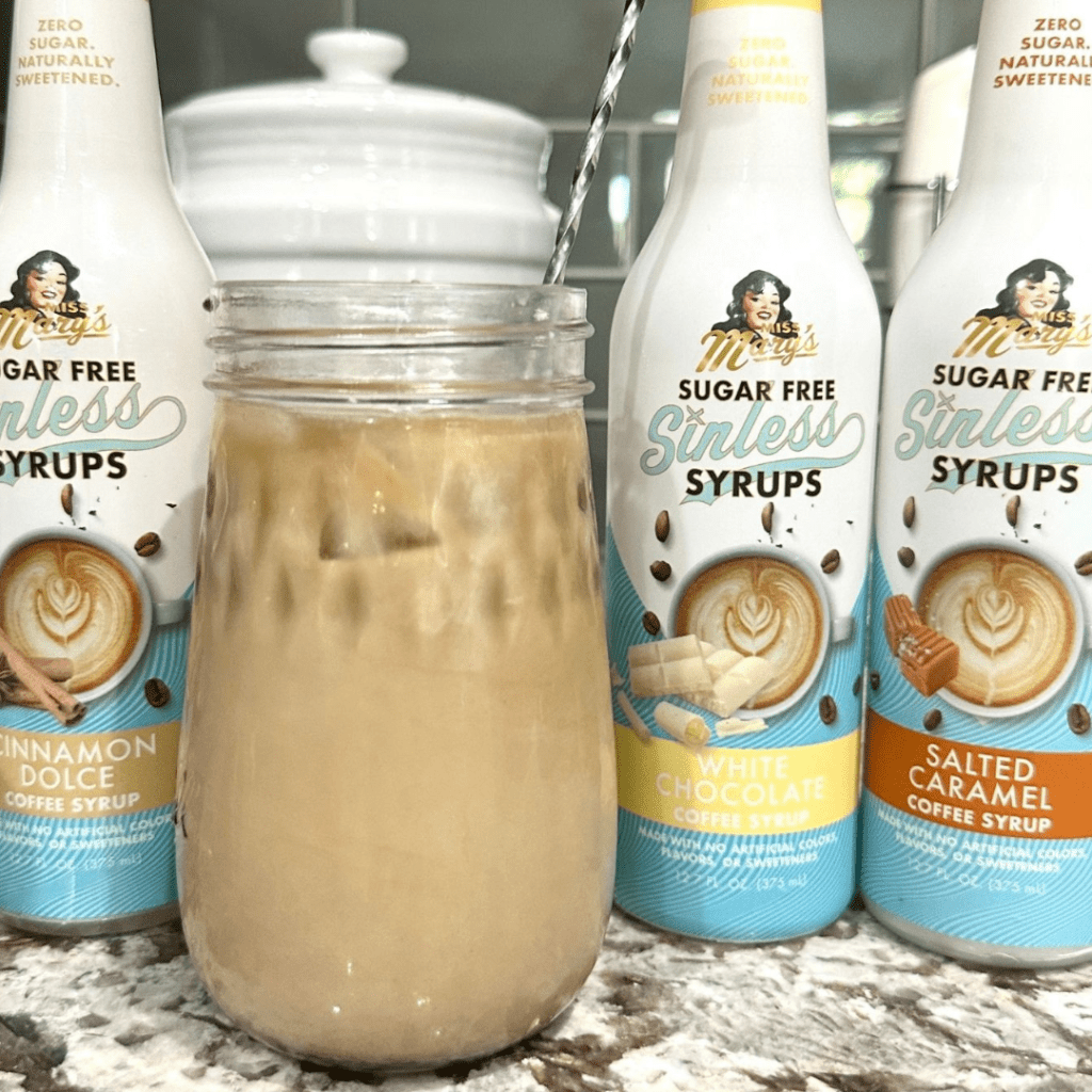An iced coffee drink sits on a countertop, surrounded by Miss Mary's Sugar Free Sinless Syrups in Cinnamon Dolce, White Chocolate, and Salted Caramel flavors.