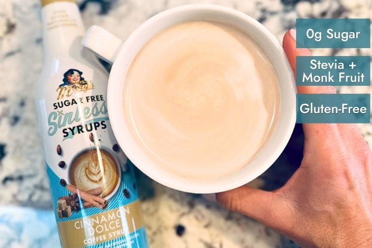 A hand holds a white ceramic mug filled with a light beige coffee drink, next to a bottle of Miss Mary's Sugar Free Sinless Syrup in Cinnamon Dolce flavor. Three teal text badges on the right read '0g Sugar', 'Stevia + Monk Fruit', and 'Gluten-Free'.