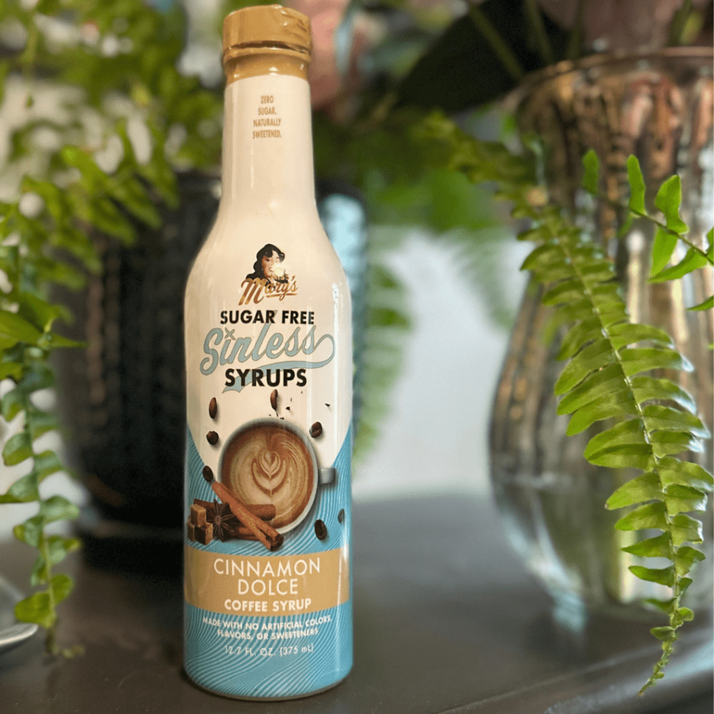 A bottle of Magic Sugar Free Stirless Syrups Cinnamon Dolce coffee syrup, standing on a dark surface with green plants in the background.