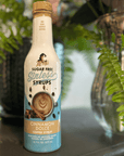 A bottle of Magic Sugar Free Stirless Syrups Cinnamon Dolce coffee syrup, standing on a dark surface with green plants in the background.