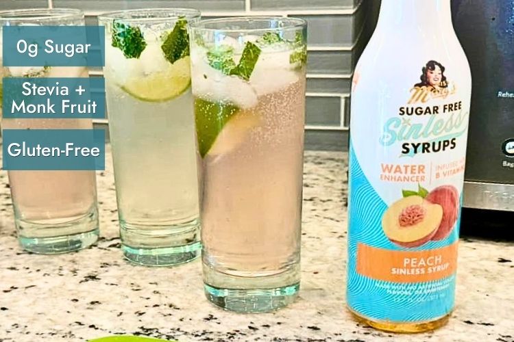 A bottle of Sugar Free Sinless Syrups Peach Sinless Syrup stands next to three glasses with text "0g sugar, stevia and monk fruit, gluten-free" on kitchen countertop.