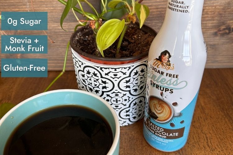 Miss Mary's Salted Dark Chocolate Sugar Free Sinless Syrup with blue coffee mug and house plant on wood background with attributes "0g Sugar, Stevia + Monk Fruit, and Gluten-Free" text