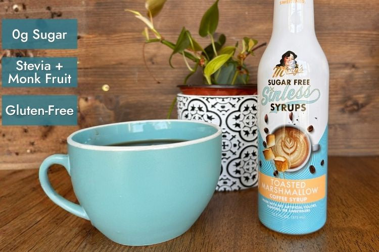 A bottle of sugar-free coffee syrup stands on a wooden table beside a light blue mug, with a small potted plant in the background. Text on the left reads '0g Sugar, Stevia + Monk Fruit, Gluten-Free'.