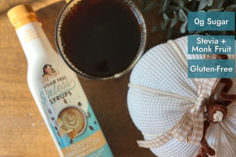 Toasted Marshmallow bottle of Sinless Syrup with coffee and pumpkin on wood table and text "0g Sugar, Stevia + Monk Fruit, Gluten-Free"