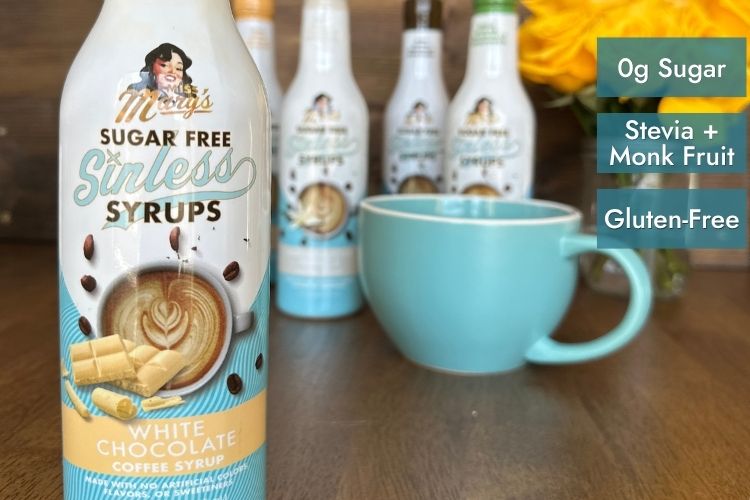 Miss Mary's Sugar Free Sinless Syrups in White Chocolate Coffee Syrup is shown in the foreground on a wooden table, with several bottles blurred in the background and a light blue mug. Text overlays on the right state '0g Sugar', 'Stevia + Monk Fruit', and 'Gluten-Free'.