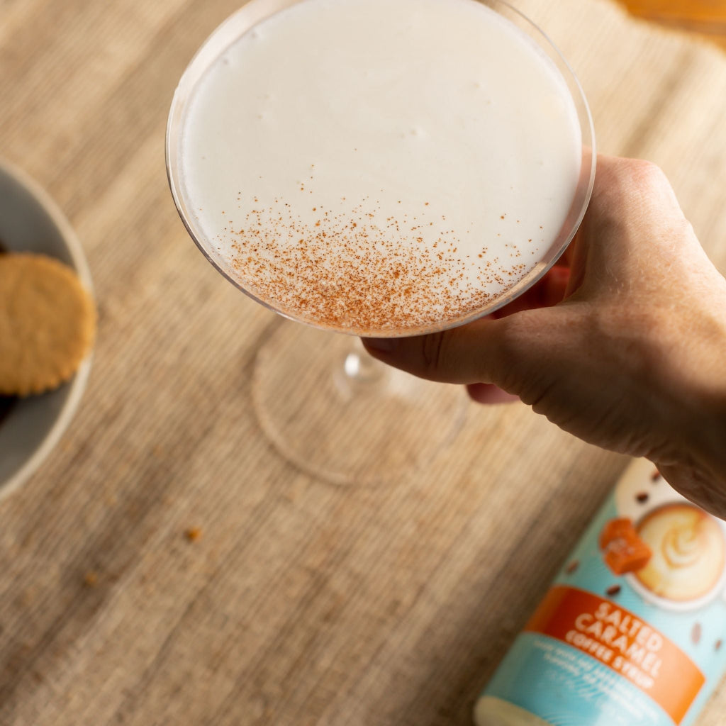 A hand holds a salted caramel martini with a dusting of cinnamon with a bottle of Salted Caramel Sinless Syrup  blurred in the background.