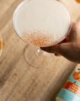 A hand holds a salted caramel martini with a dusting of cinnamon with a bottle of Salted Caramel Sinless Syrup  blurred in the background.