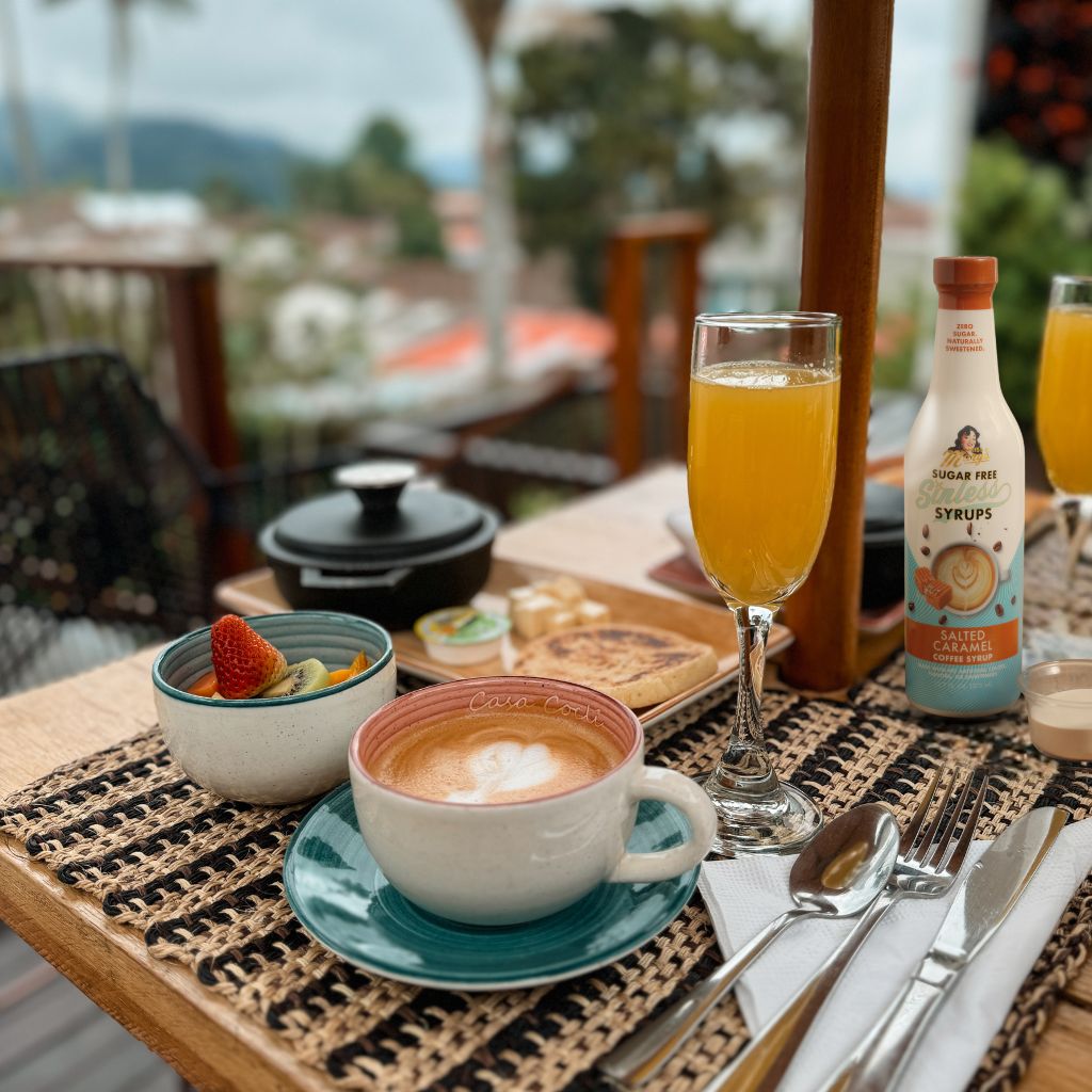 Breakfast setting with coffee, fruit, orange juice, and salted caramel Sinless Syrup on a table with a blurred outdoor background.