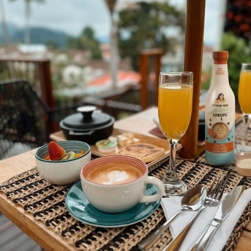 A styled breakfast table scene with coffee, orange juice, a bottle of sugar-free Salted Caramel syrup, fruit, toast, and utensils on a woven placemat, with an outdoor balcony and blurred background.