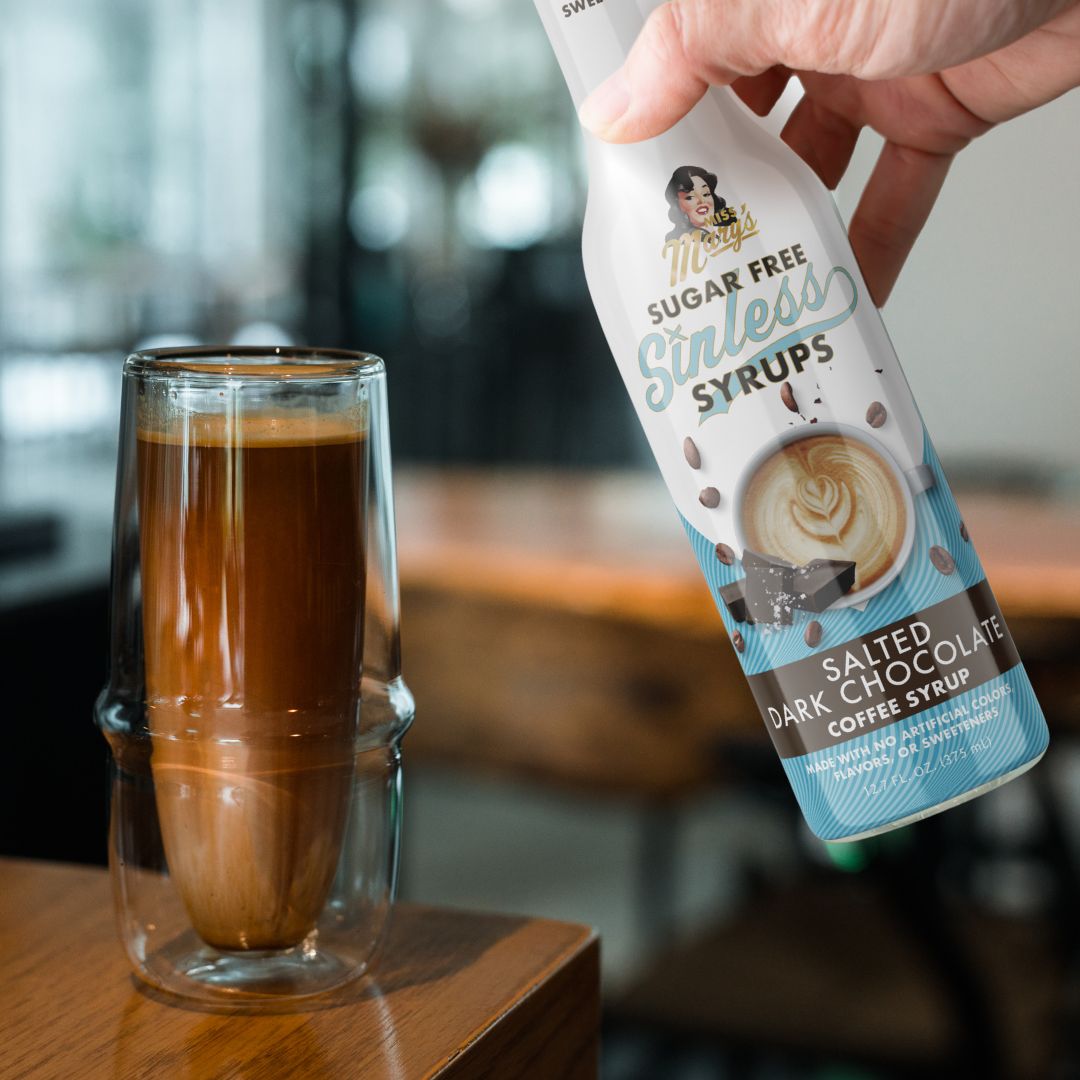 Hand holding a bottle of sugar-free salted dark chocolate syrup next to a tall espresso drink.