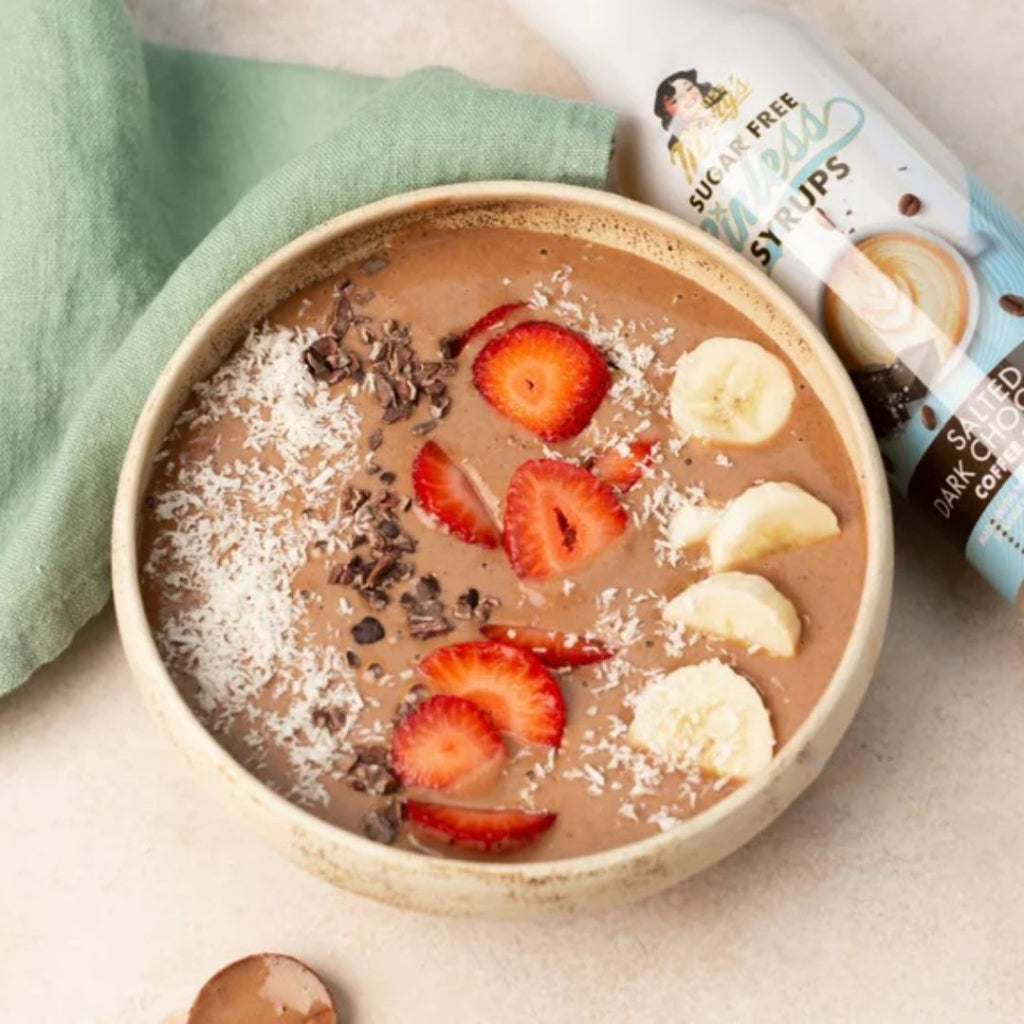A chocolate smoothie bowl with sliced strawberries, banana, coconut flakes, and chocolate chips, styled with a green cloth and Miss Mary's Salted Dark Chocolate Sinless Syrup