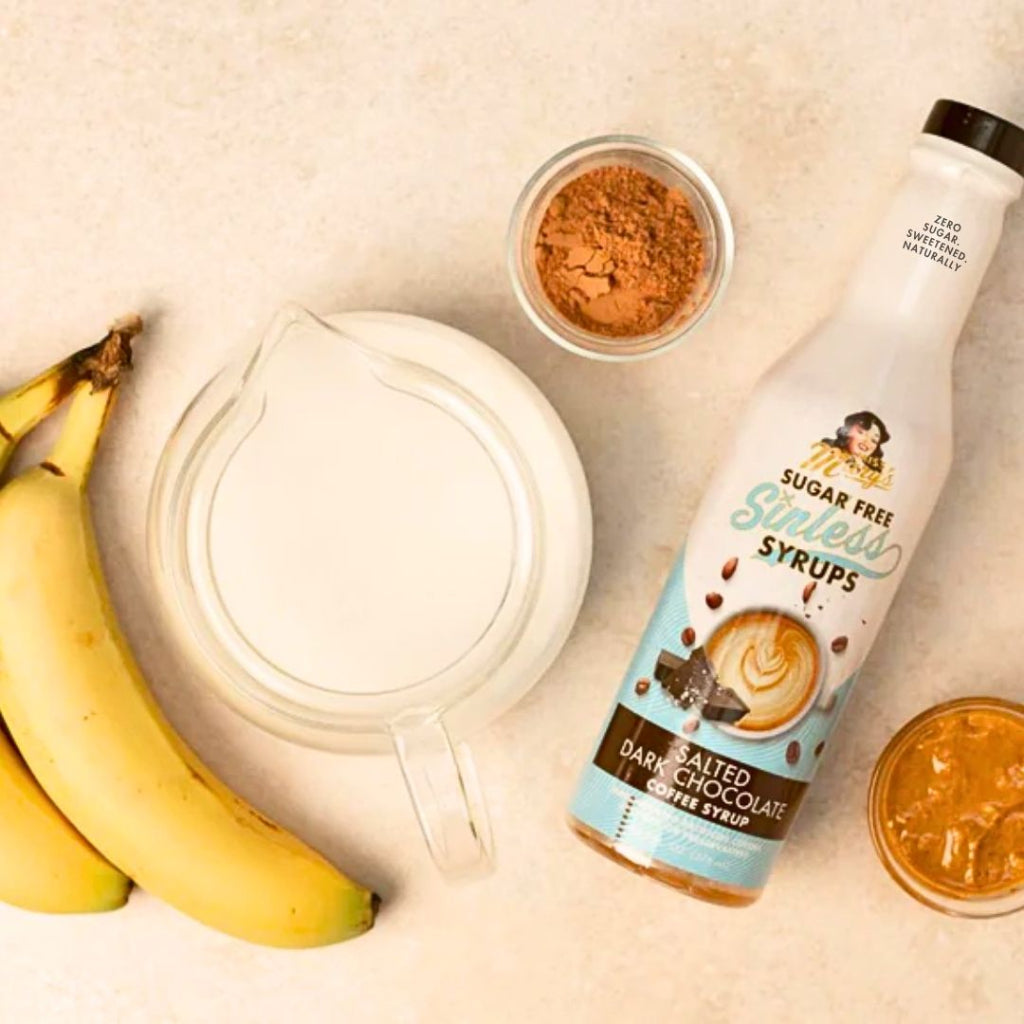 A bottle of sugar-free sweetened syrups labeled coffee syrup, arranged on a light beige surface with bananas, a glass pitcher, cocoa powder, and a small jar.