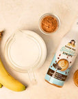 A bottle of sugar-free sweetened syrups labeled coffee syrup, arranged on a light beige surface with bananas, a glass pitcher, cocoa powder, and a small jar.