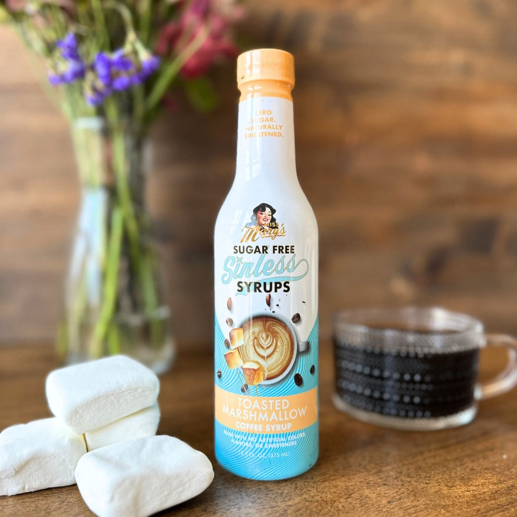 Toasted Marshmallow Sinless Syrup bottle with large marshmallows on a wood surface with coffee and flowers blurred in the background