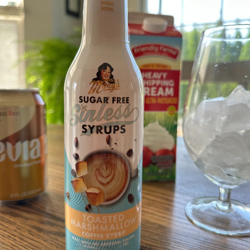 Bottle of Mrs. Mary's Sugar Free Sinless Syrups Toasted Marshmallow flavor on a table with a can, carton, and glass.