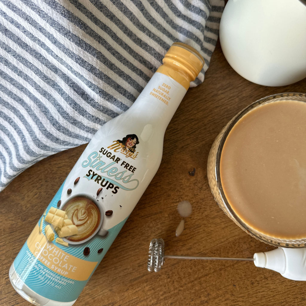 White Chocolate Sinless Syrup on wood table with frother and latte, striped towel.