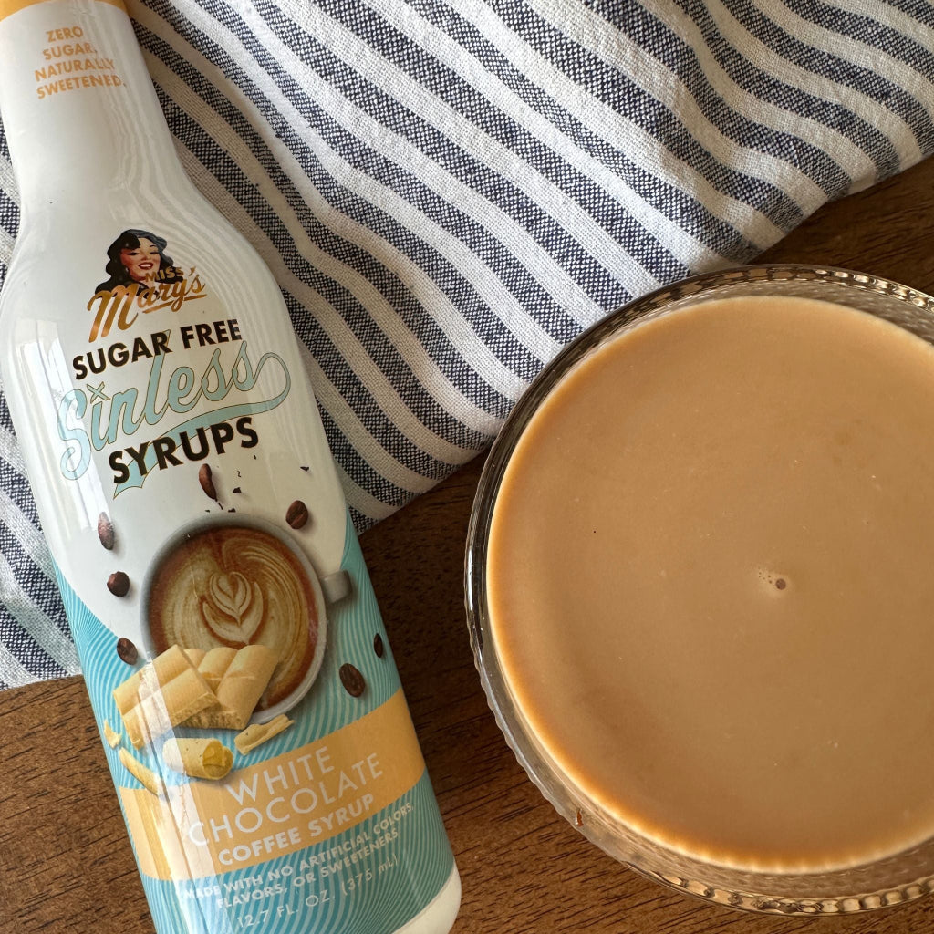 A bottle of Mary's Sugar Free Sinless Syrups White Chocolate Coffee Syrup next to a glass filled with the light brown coffee syrup, set on a wooden table with a striped cloth background.